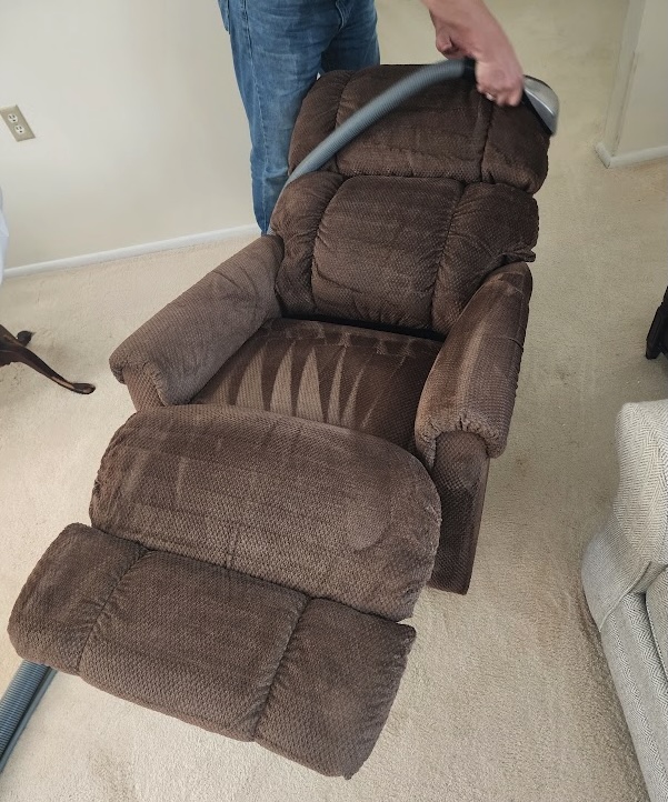 Technician using a vacuum extraction tool to clean a brown upholstered recliner in a residential living room.